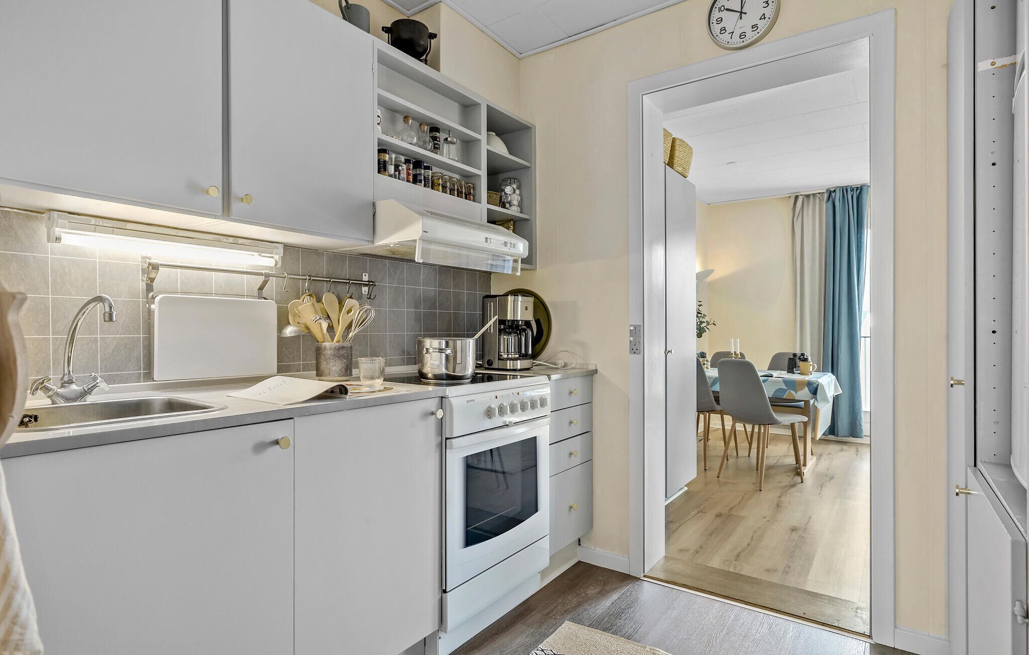 A modern kitchen with white cabinets, a built-in oven, and a coffee maker. There is a dining area visible through an open door.