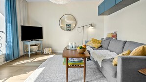 Un salon moderne avec un téléviseur à écran plat, un miroir rond, une table d’appoint sur laquelle se trouve un saladier rempli de fruits, et un canapé gris agrémenté de coussins jaunes.