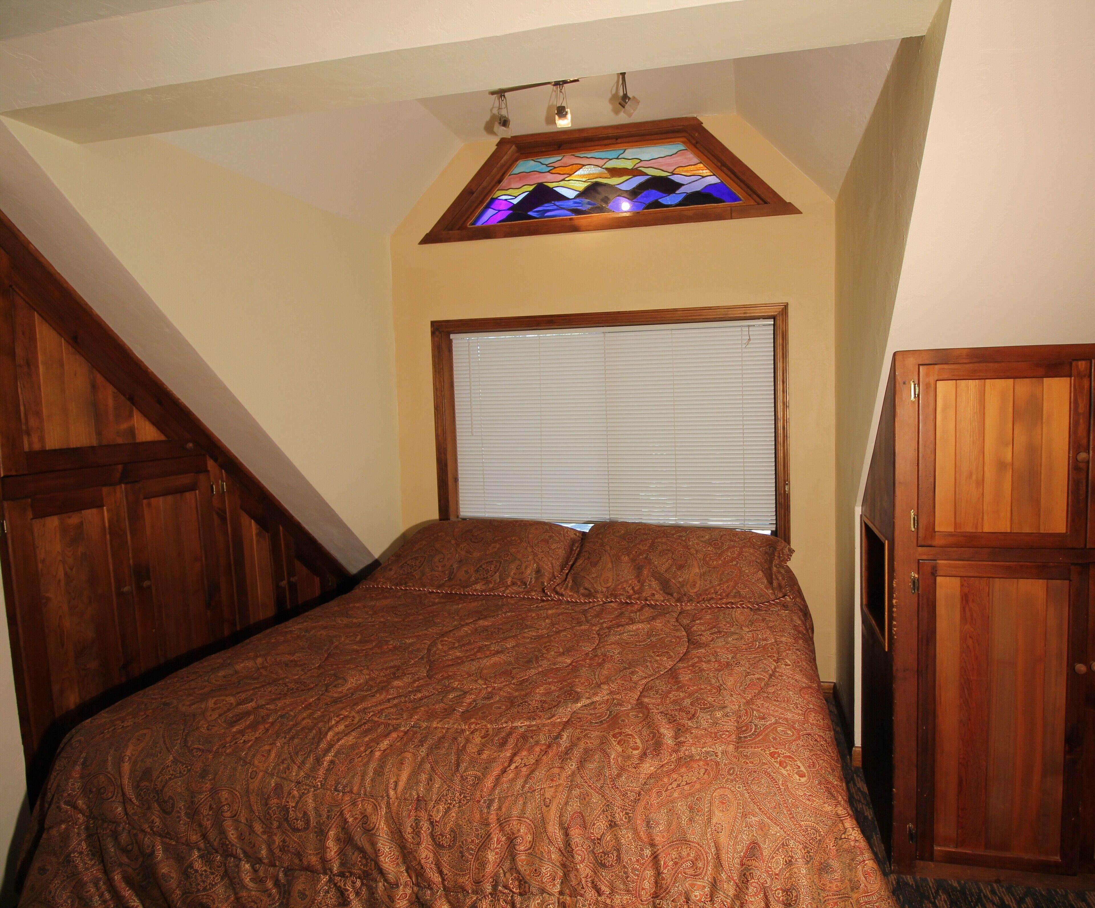 A bedroom with a bed, a window with blinds, a wooden headboard, and a stained glass window above.
