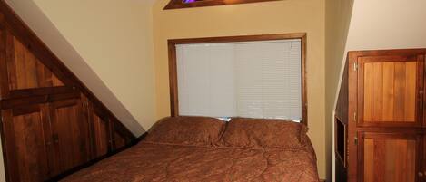 A bedroom with a bed, a window with blinds, a wooden headboard, and a stained glass window above.