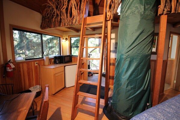 L’intérieur de la cabane comprend une échelle en bois, un coin cuisine, un espace repas et de grandes fenêtres donnant sur la forêt.