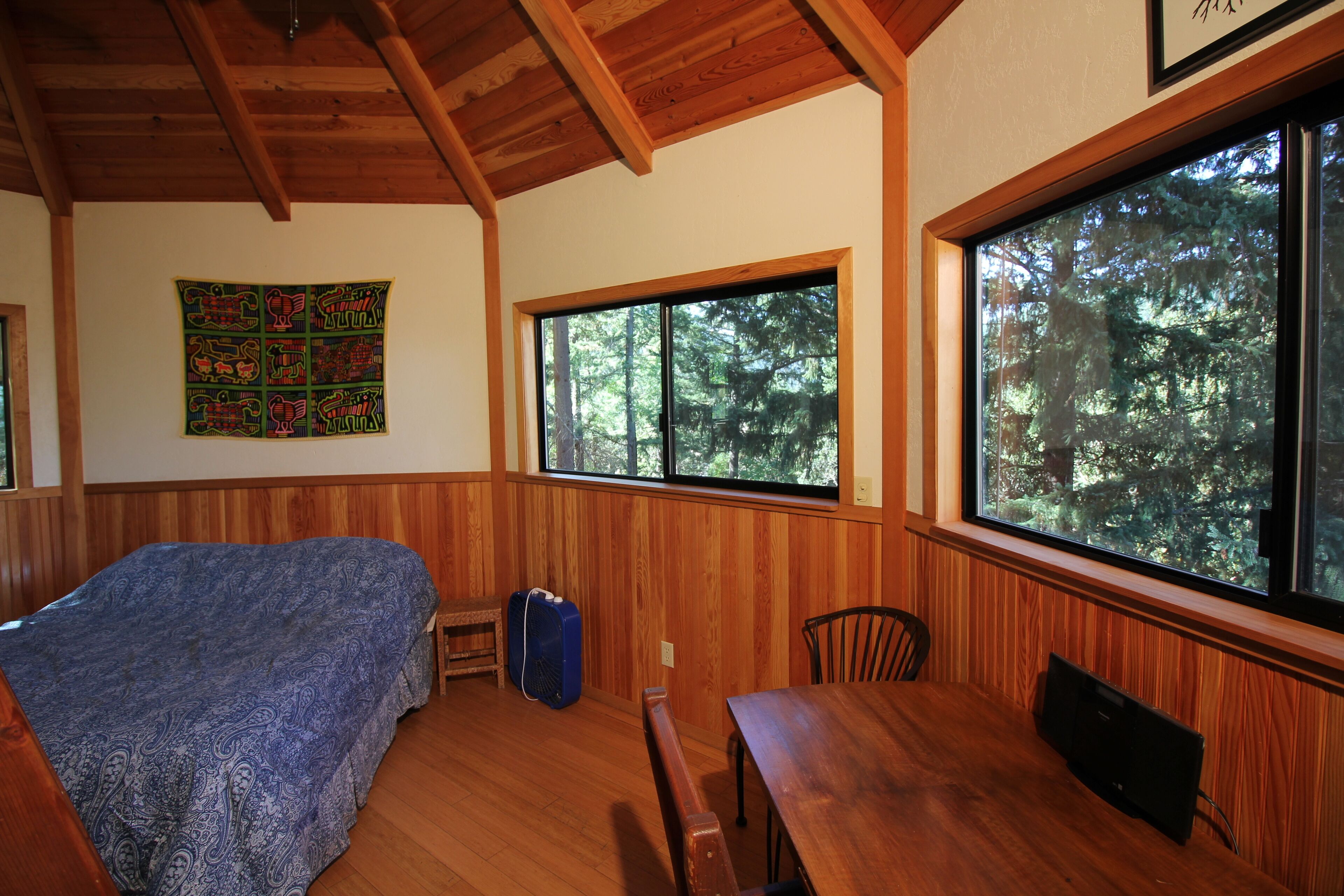 Una habitación con una cama, una mesa de madera y ventanas amplias con vistas al bosque.