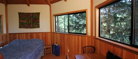 Una habitación con una cama, una mesa de madera y ventanas amplias con vistas al bosque.
