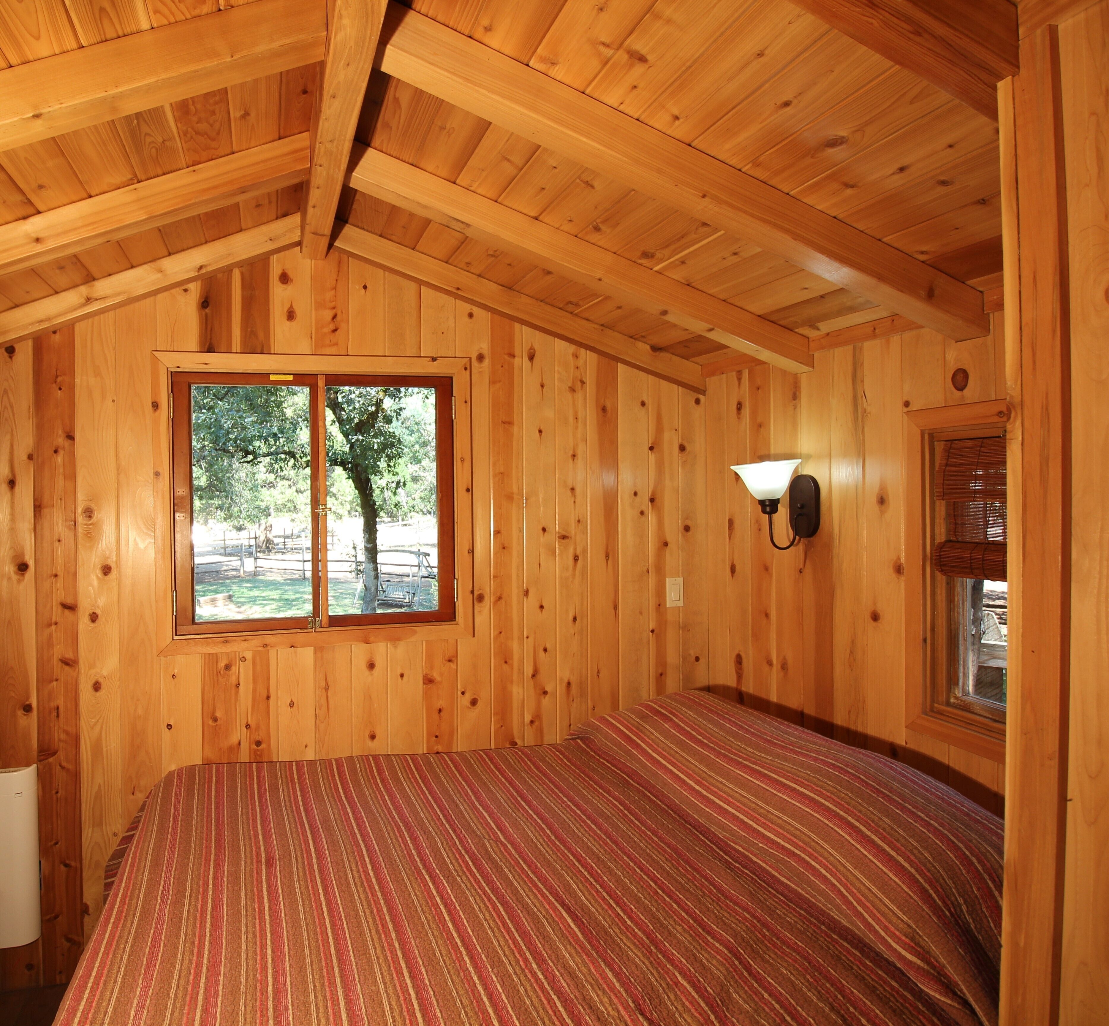 A wooden cabin room with a bed, a window with a view of trees, and a wall-mounted light fixture.