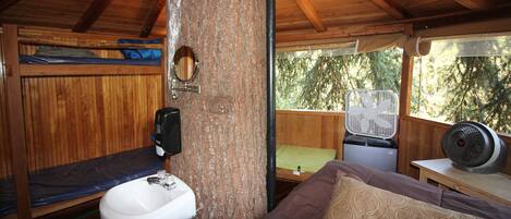 A wooden cabin interior with bunk beds, a sink, a fan, and a window overlooking trees.