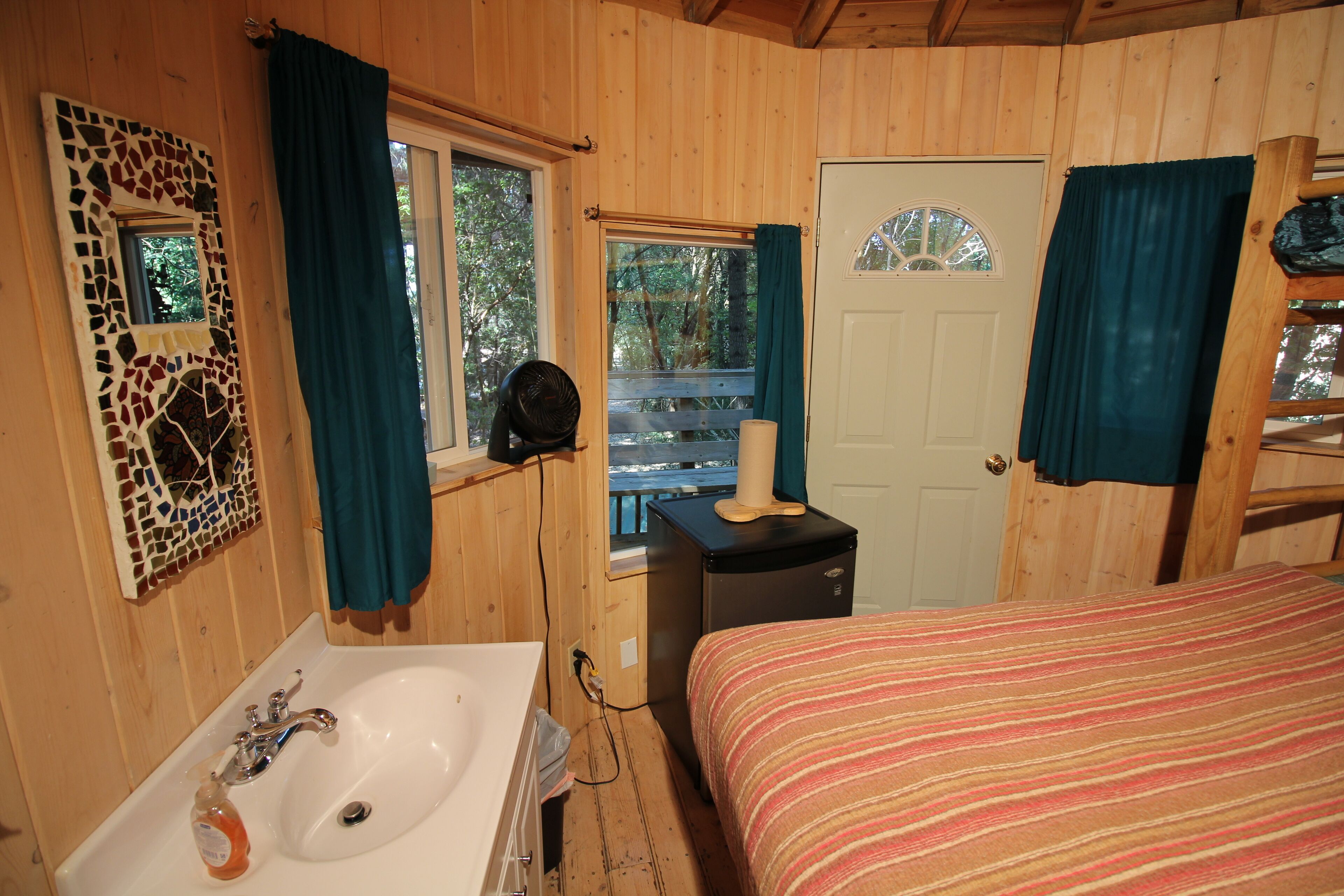 A wooden cabin bedroom with a bed, a sink, a mirror, and a window with curtains.