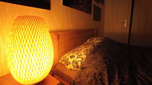 A bedroom with a bed, a patterned blanket, a wall-mounted lamp, and a door. - Le Cocon Urbain" 2-room apartment (Neuilly-Plaisance)
