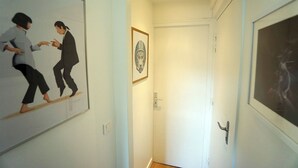 A hallway with a framed picture of two people on the left, a framed picture above a door on the right, and a door with a handle. - Le Cocon Urbain" 2-room apartment (Neuilly-Plaisance)
