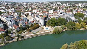 An aerial view of a city with a river, featuring residential buildings, a bridge, and a swimming area. - Le Cocon Urbain" 2-room apartment (Neuilly-Plaisance)
