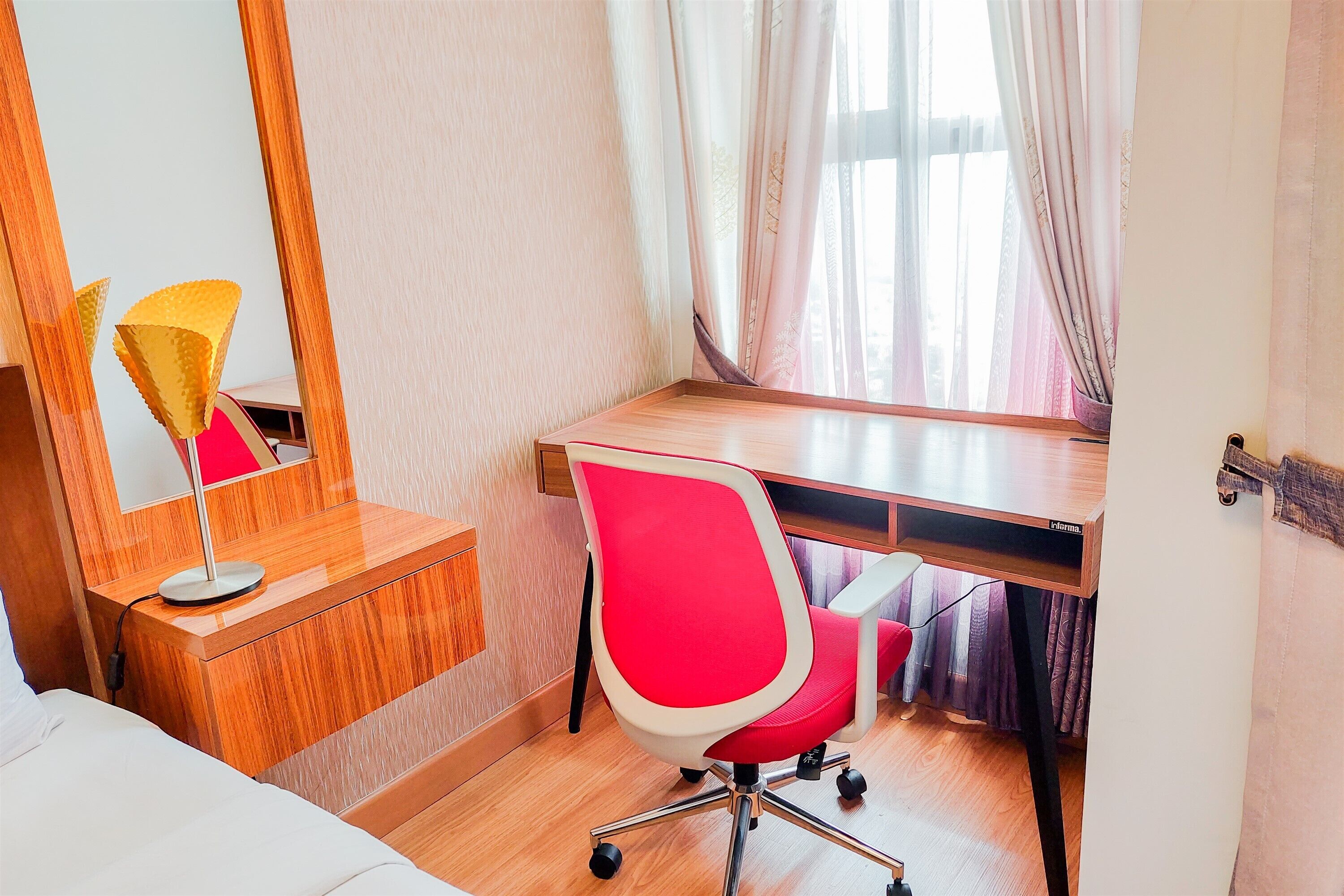 A hotel room with a wooden desk, a red office chair, a bedside table with a lamp, and a large mirror.