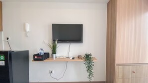 Camera d'albergo con TV a schermo piatto fissata al muro, un minibar, una bottiglia d'acqua, una pianta in vaso e un mobiletto in legno.