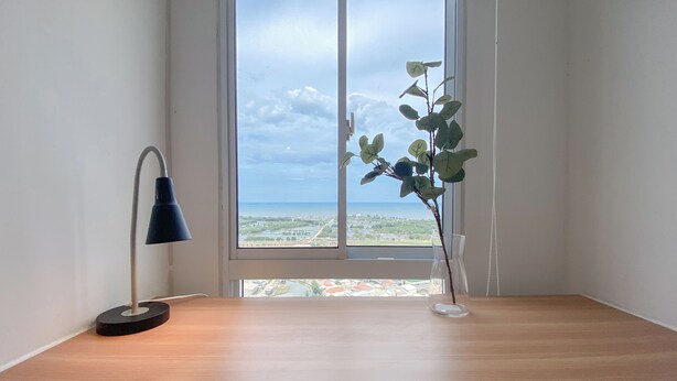 Una stanza con una scrivania in legno, una lampada da tavolo nera e una pianta in vaso vicino a una finestra che offre vista su uno specchio d'acqua e vegetazione.