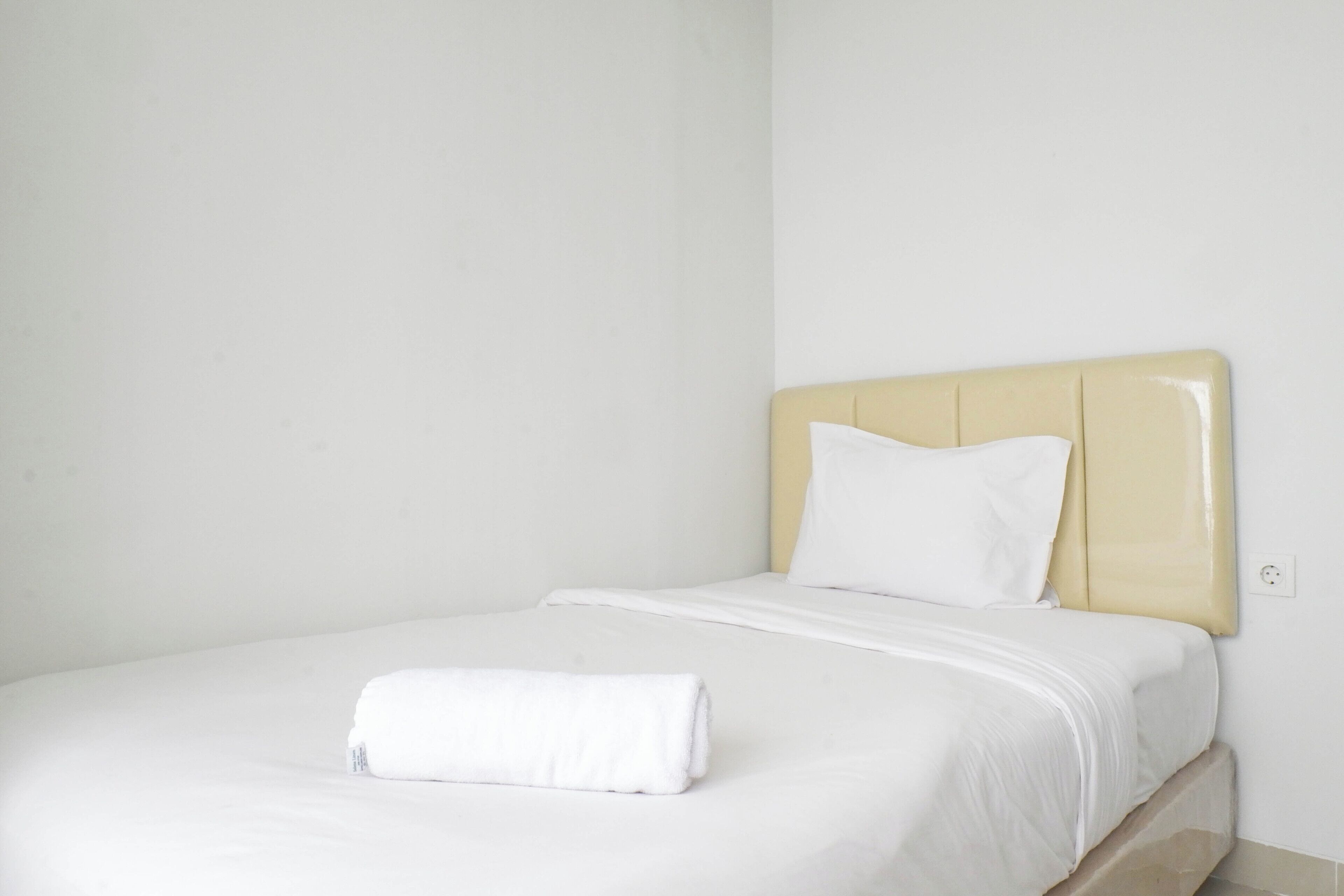 Een eenpersoonsbed met een beige hoofdeinde en witte beddengoed, een opgevouwen witte handdoek op het bed, en een effen witte muur.