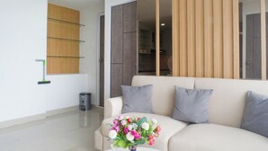 A modern living room with a beige sofa, grey cushions, and a vase of flowers. There is a wooden panel wall and a glass shelf unit. - Exquisite And Vibrant 2Br Apartment At Taman Melati Surabaya (Surabaya)