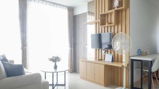 A modern living room with a sofa, a small round table with a vase, a TV on a wooden stand, a standing fan, and a desk with a chair. - Exquisite And Vibrant 2Br Apartment At Taman Melati Surabaya (Surabaya)