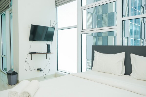 A modern hotel room with a large bed, a flat-screen TV mounted on the wall, and large windows offering a view of the cityscape.