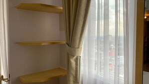 A room with a window, a curtain, and three wooden shelves against the wall. - Cozy Nice Studio At Signature Park Grande Apartment (Jakarta)