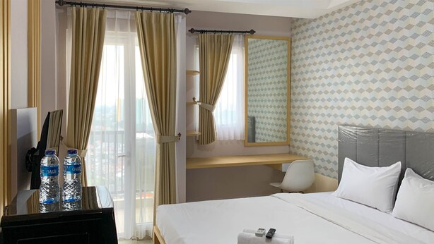 A hotel room with a bed, a desk with a chair, a mirror, and two bottles of water. - Cozy Nice Studio At Signature Park Grande Apartment (Jakarta)