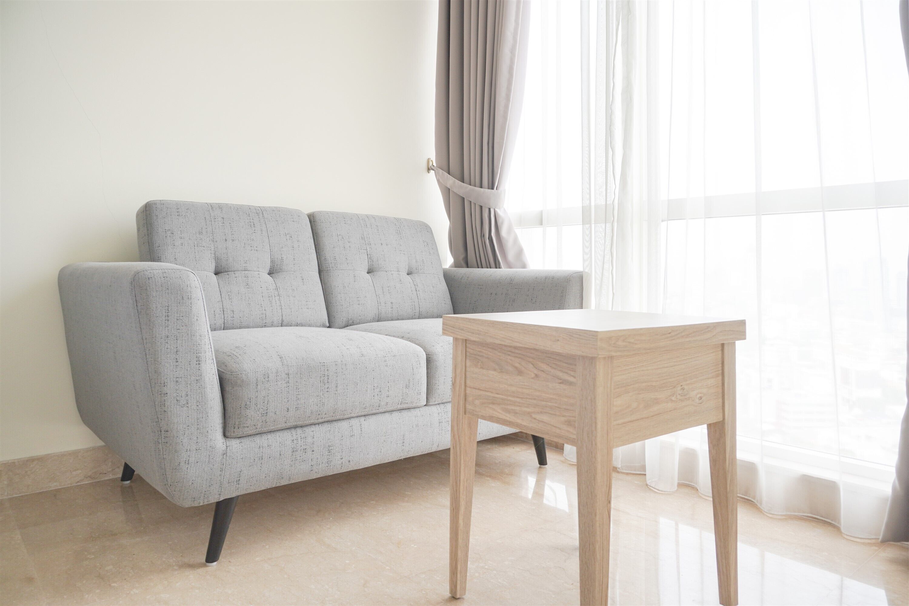 Una sala moderna con un sofá gris, una mesa de centro de madera y cortinas translúcidas en una ventana.