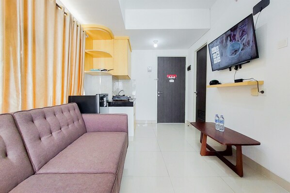 A modern living room with a sofa, a TV mounted on the wall, a wooden table with water bottles, and a kitchen area in the background. - Nice And Cozy Living 2Br Serpong Garden Apartment (Cisauk)