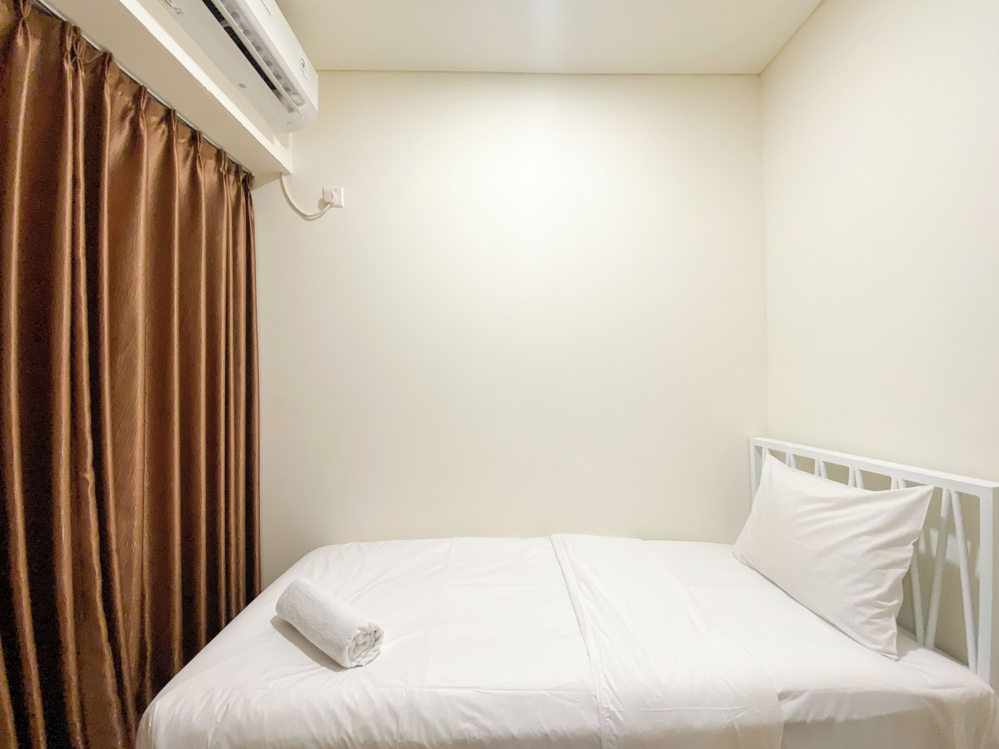 A small, neatly made bed with white bedding and a rolled white towel, a white headboard, and a brown curtain.