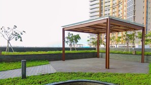 Un espace extérieur abrité, doté d’une structure en bois, entouré de verdure et bordé d’un chemin pavé.