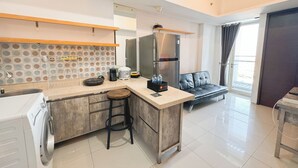 Una cocina moderna con lavadora incorporada, un refrigerador y una cafetera. Hay un comedor con un sofá de cuero negro y una ventana con cortinas.
