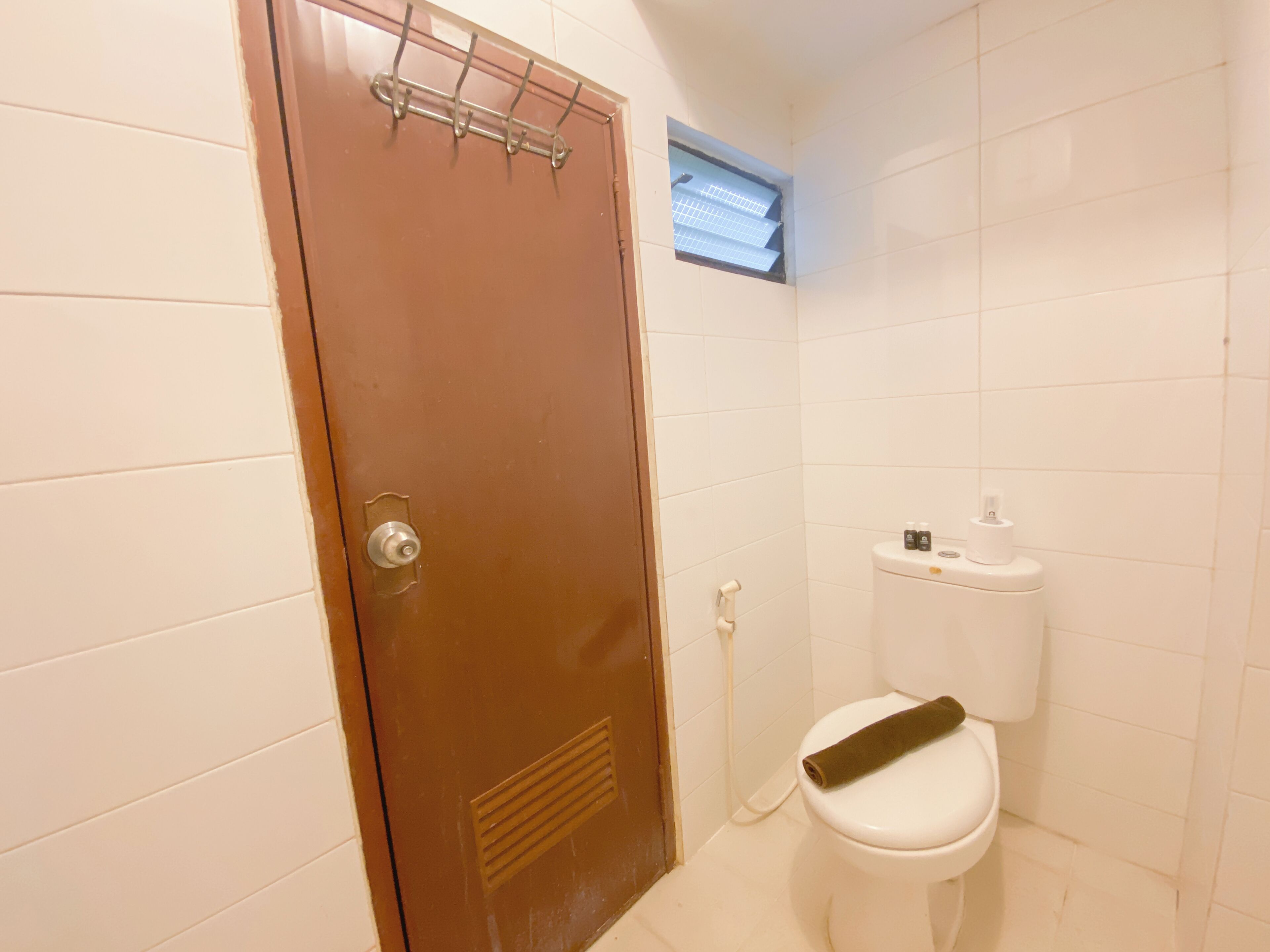 Une salle de bain avec des toilettes blanches, une serviette posée sur la cuvette, une fenêtre au-dessus des toilettes et un porte-serviettes sur la porte.