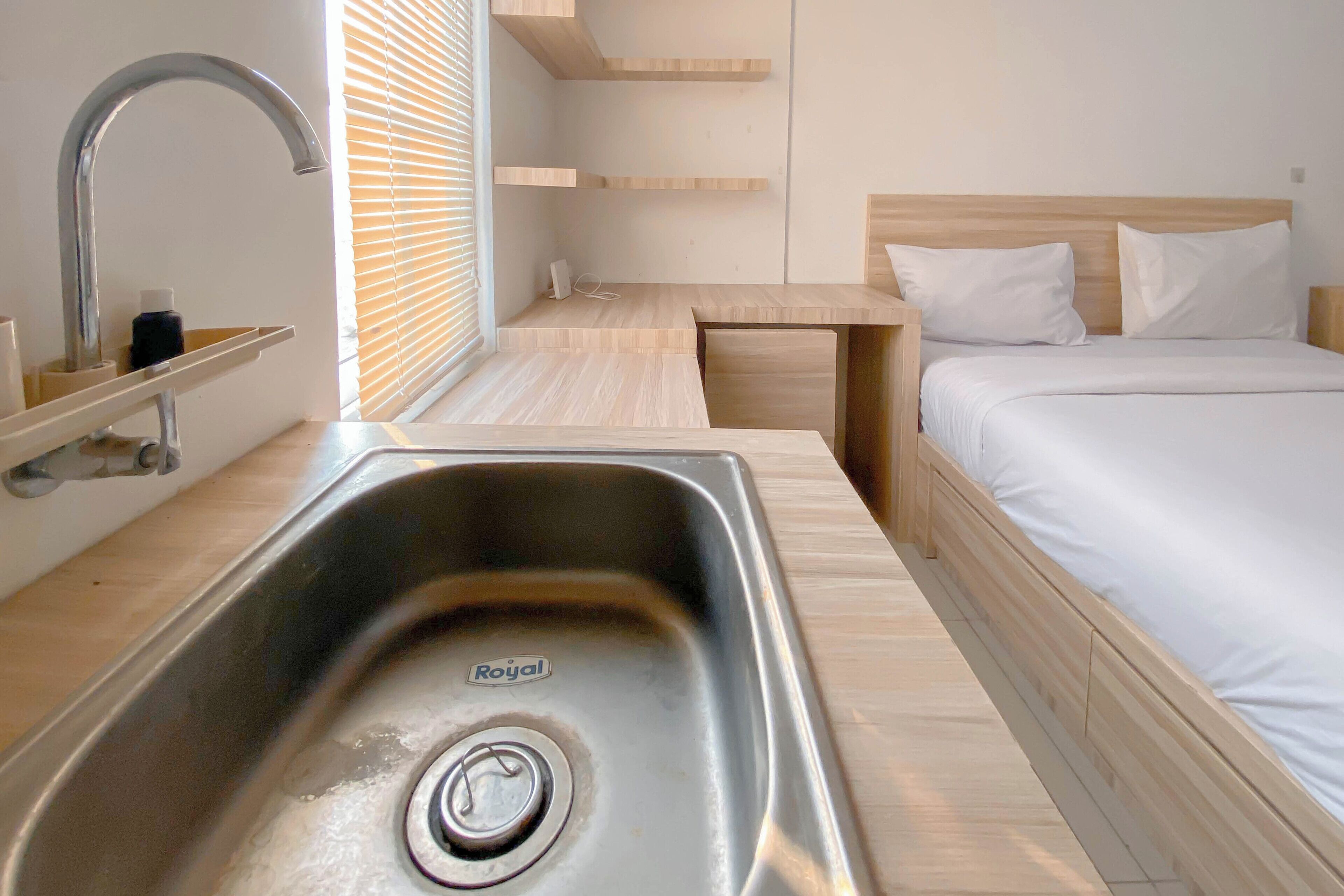Een moderne hotelkamer met een inbouwspoelbak, houten nachtkastjes, een bed met witte lakens en een raam met luxaflex.