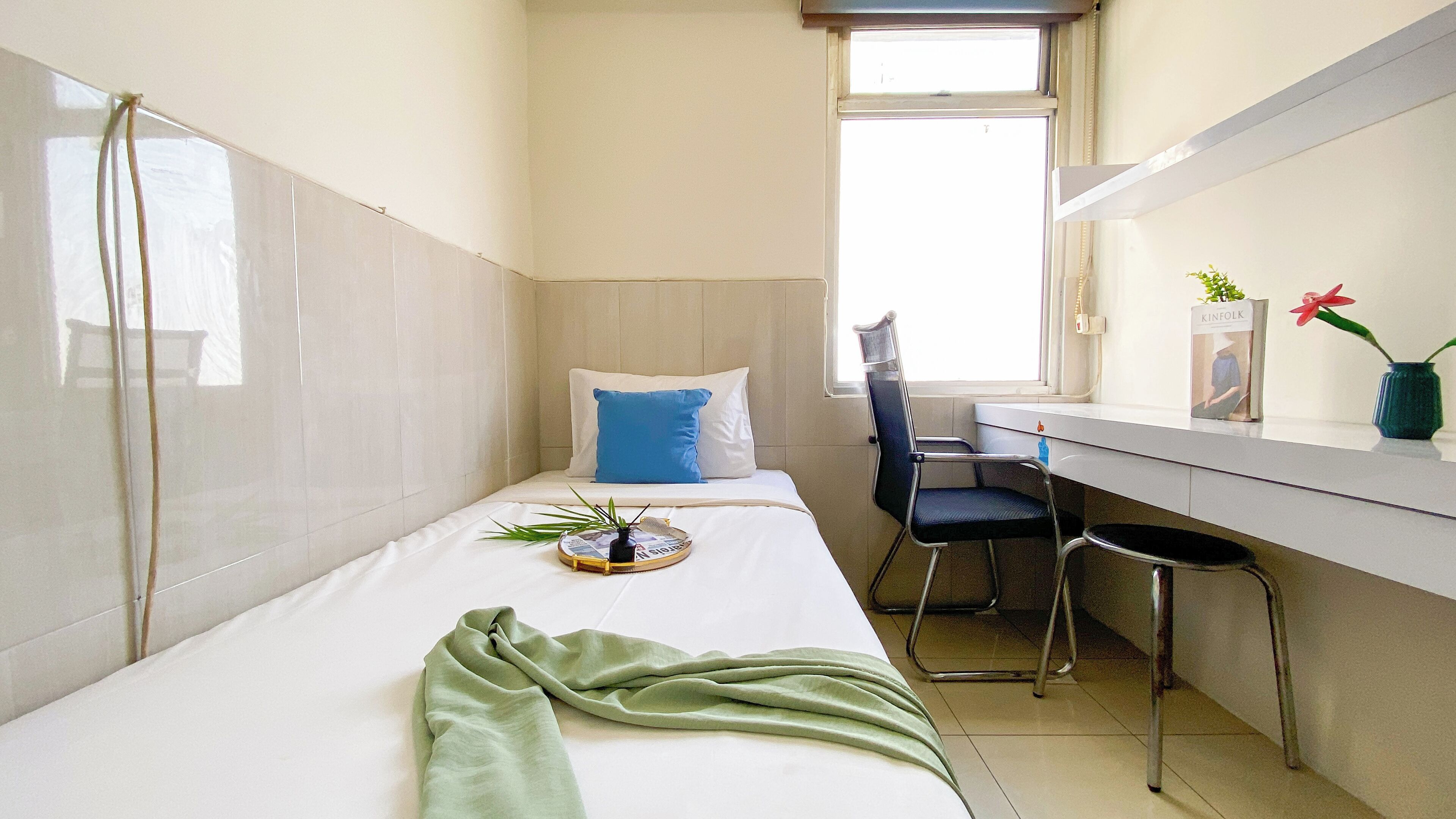 A small, compact room with a single bed, a desk, and a chair. There is a window providing natural light, and a small plant on the desk.