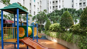 A playground with slides and climbing structures, surrounded by greenery and a building in the background. - Spacious And Homey 3Br At Green Bay Pluit Apartment (Jakarta)
