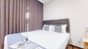 A hotel room with a large bed, a bedside table, a nightstand, a window with curtains, and a wall-mounted outlet. - Good Place And Homey 3Br At Asatti Apartment (Pagedangan)