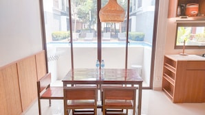 Een eethoek met een houten tafel en stoelen, een geweven hanglamp, en uitzicht op een zwembad en groen buiten.