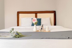 A neatly made bed with white linens, a wooden headboard, a teal pillow, a white vase with a pink flower, and a small table with a glass vase and a candle. - Best Choice And Modern 2Br Green Pramuka City Apartment (Jakarta)