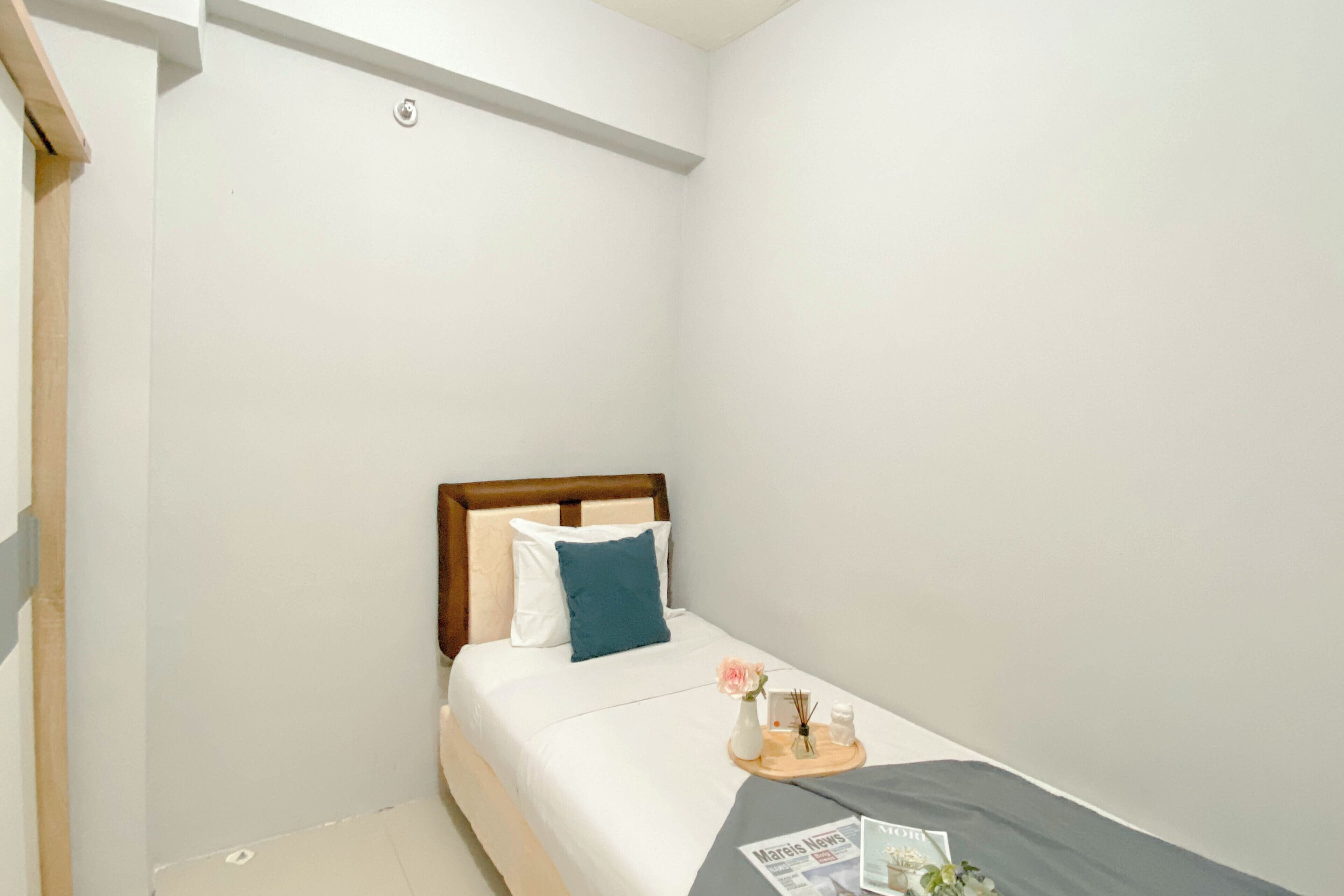 Una habitación pequeña y limpia con una cama, cabecera de madera, mesita de noche con lámpara, jarrón con flores y bandeja con periódico.