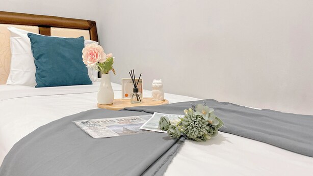 A neatly made bed with a grey blanket, a teal pillow, and a white pillow. There is a bouquet of flowers, a small white cat figurine, and a tray with a candle and reed diffuser on the bed. - Best Choice And Modern 2Br Green Pramuka City Apartment (Jakarta)