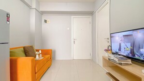A modern living room with a bright orange sofa, a flat-screen TV, and a wooden side table with a book and a vase. - Best Choice And Modern 2Br Green Pramuka City Apartment (Jakarta)