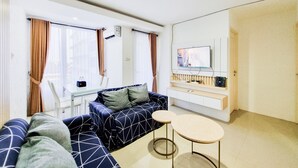 A modern living room with a sofa, a small round table, a TV mounted on the wall, and a window with curtains. - Good Place And Homey 2Br At Green Park Yogyakarta Apartment (Depok)