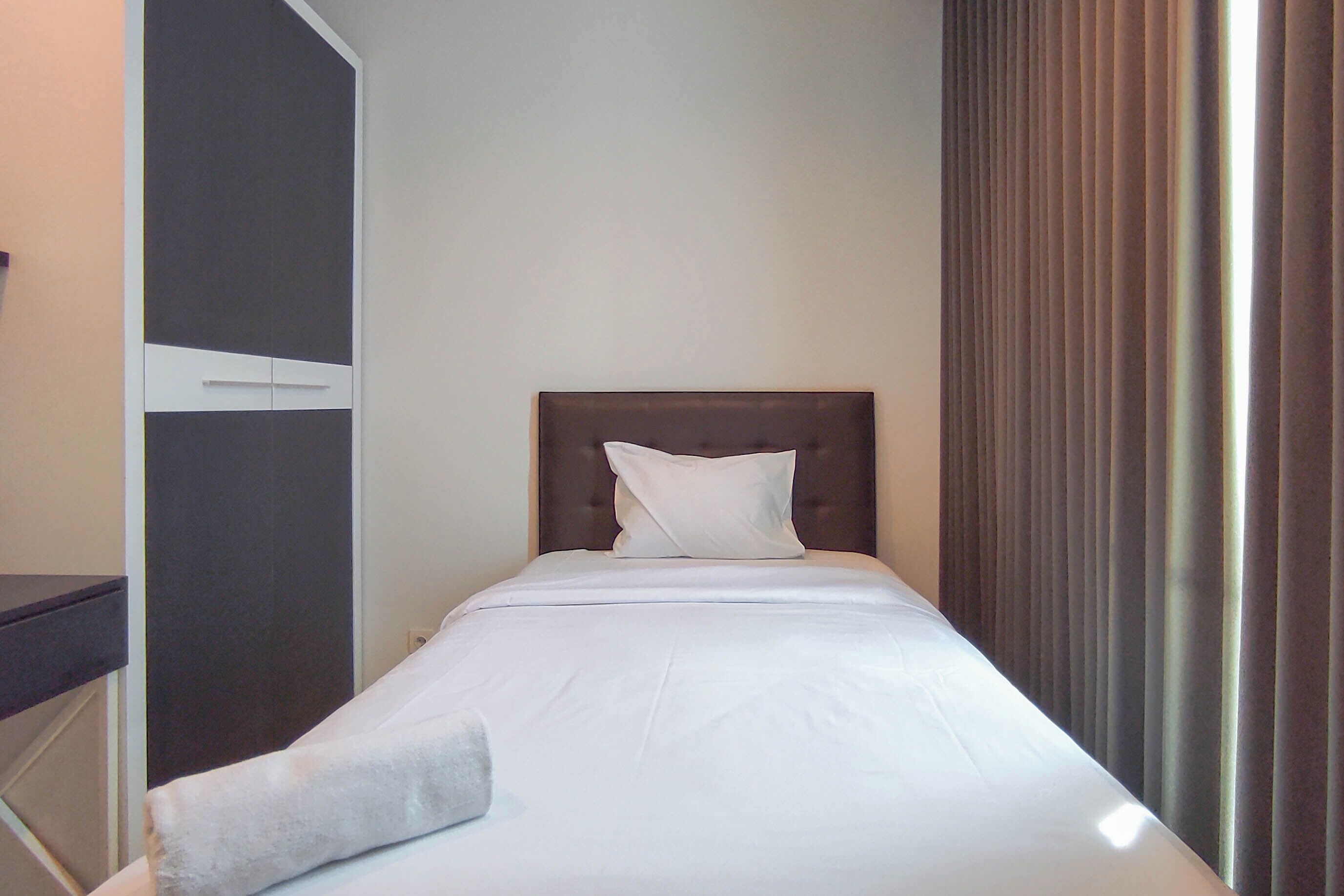 Quarto de hotel com uma cama de solteiro, cabeceira de madeira, travesseiro branco e uma mesinha de cabeceira.
