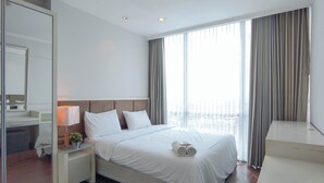 Habitación de hotel con una cama grande, una mesita de noche con lámpara, un espejo y vistas a la ciudad a través de amplios ventanales.