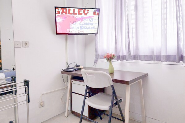Una habitación con escritorio, silla, televisor montado en la pared y ventana con cortinas.