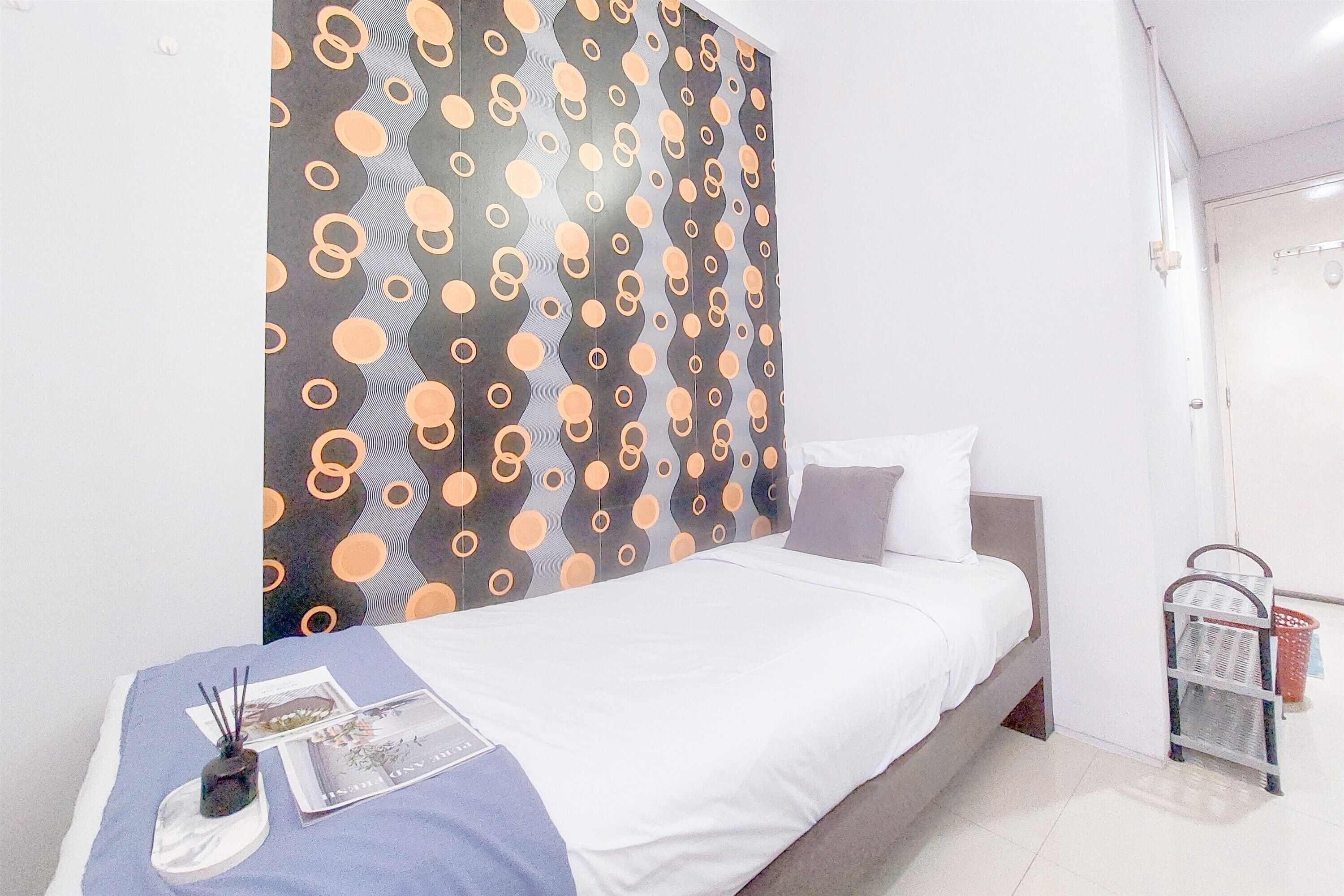Una cama con colchón blanco y cabecera gris, una mesita de noche con lámpara, y una pared con diseño circular.