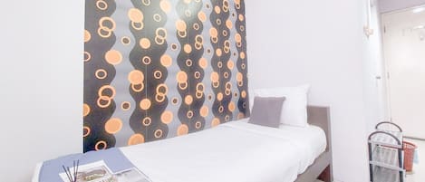 A bed with a white mattress and grey headboard, a bedside table with a lamp, and a wall with a circular pattern.
