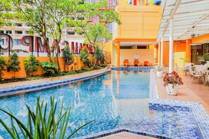 Una piscina con borde de azulejos, rodeada de vegetación y un edificio de fachada amarilla.