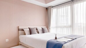 A neatly made bed with a blue blanket, a bedside table with a lamp, and sheer curtains on a window. - Comfy And Homey Studio At Casa De Parco Apartment (Pagedangan)