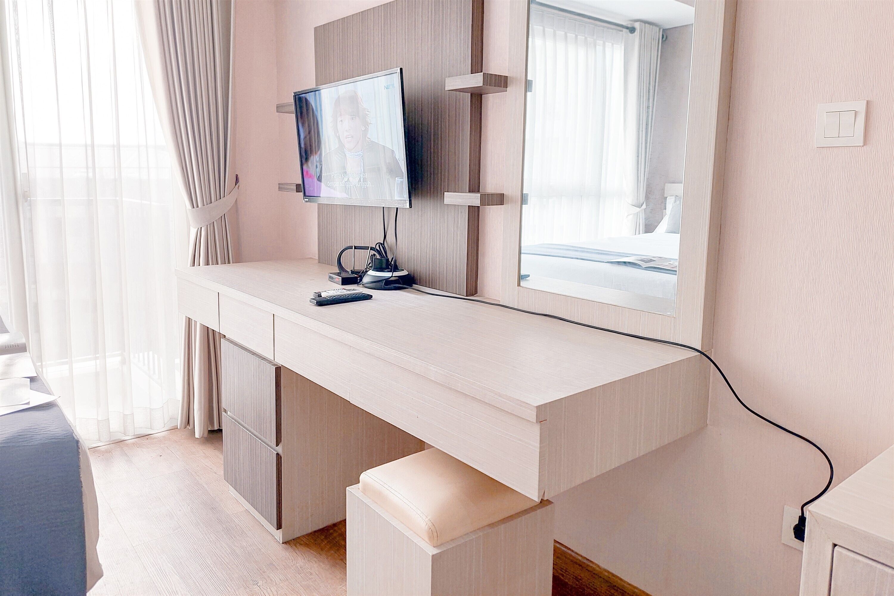 A modern hotel room with a light-colored desk, a flat-screen TV, a large mirror, and a window with sheer curtains.
