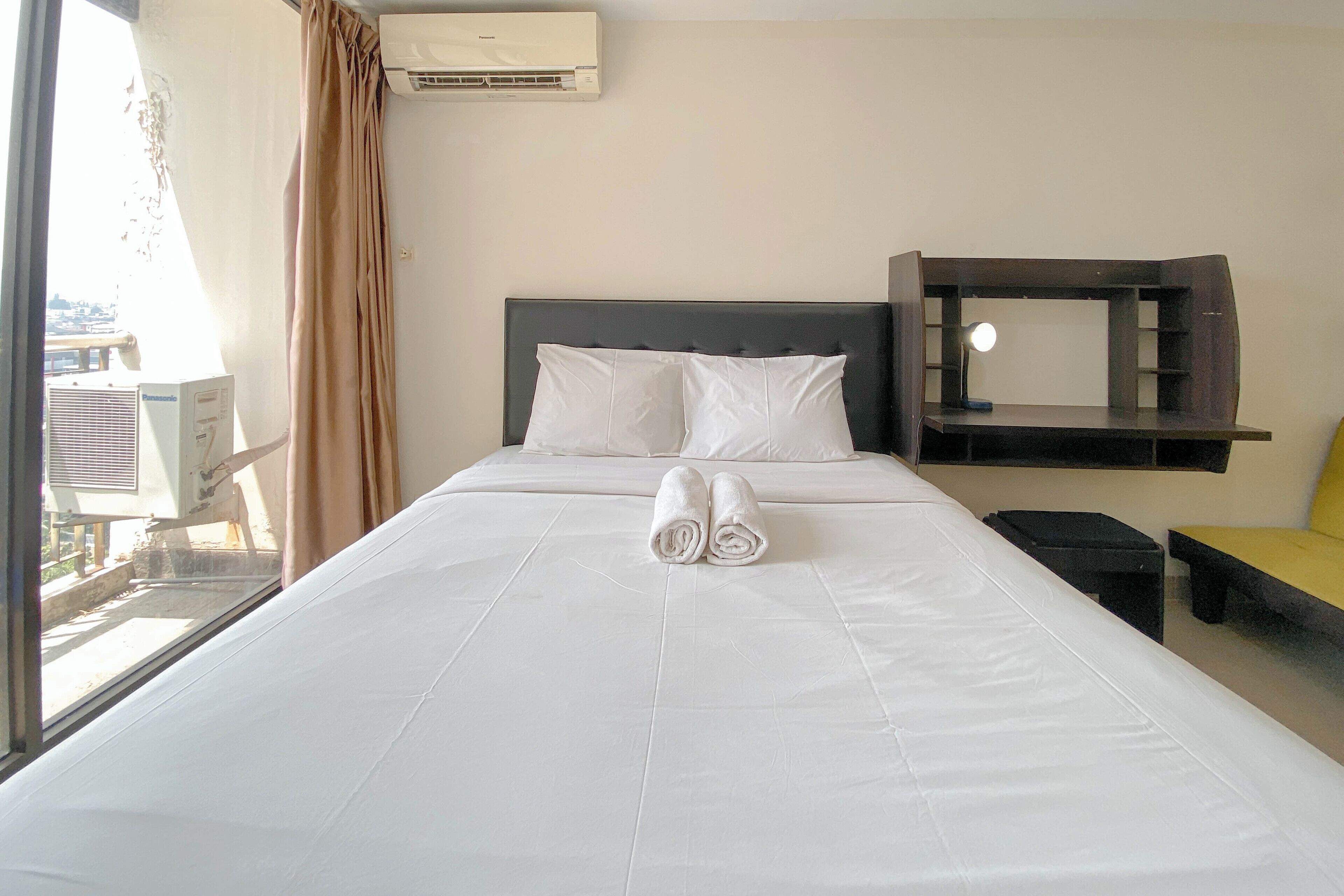 A neatly made bed with white linens and rolled towels, a bedside table, a small black stool, a yellow bench, and a wall-mounted shelf with a mirror.