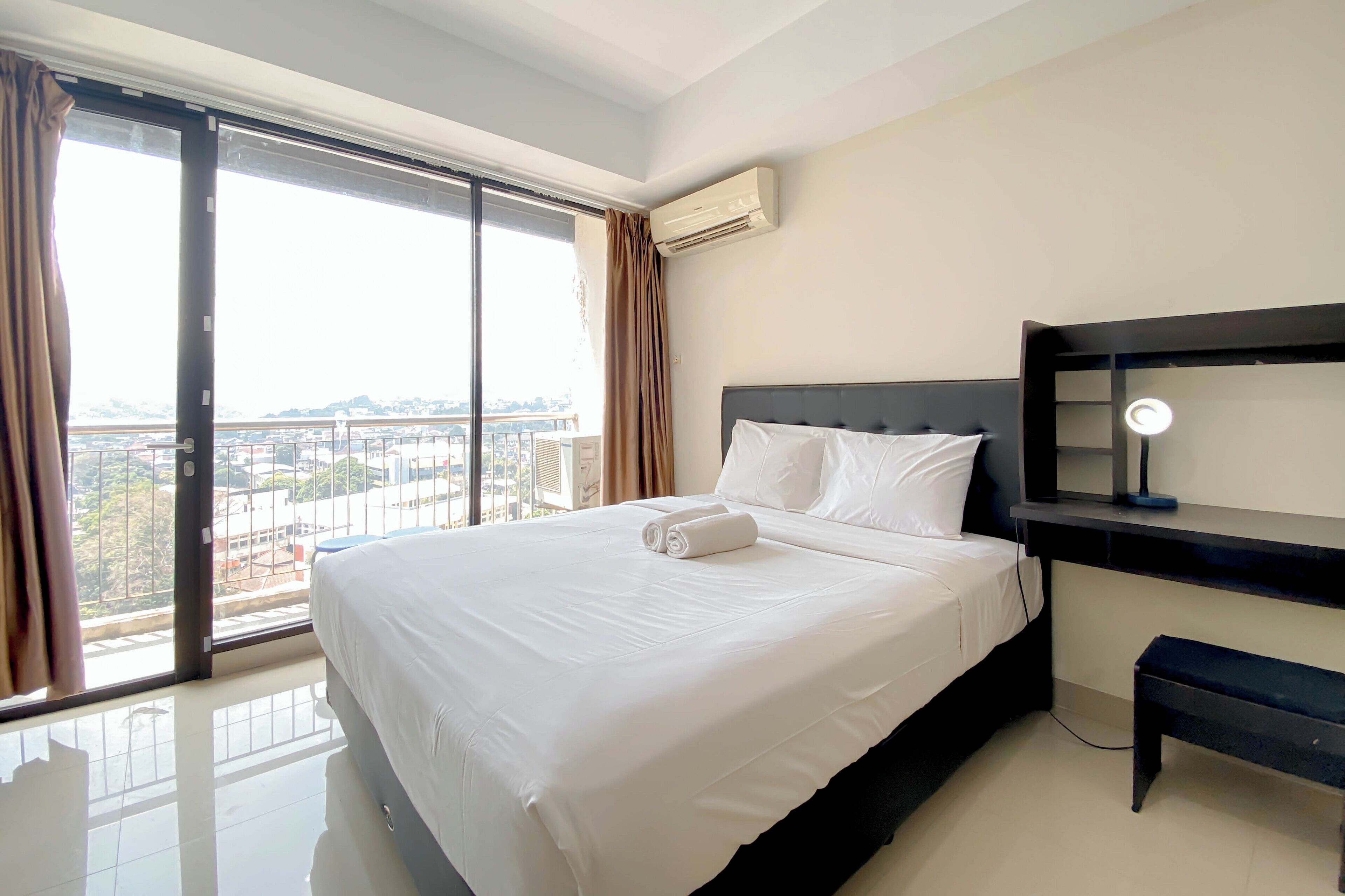 Habitación de hotel con una cama grande, una mesita de noche con lámpara y vistas a la ciudad a través de un amplio ventanal.