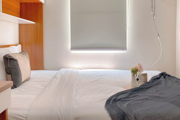 A neatly made bed with white linens, a bedside table with a vase of flowers, and a window with a white blind. - Strategic And Restful 2Br Springlake Summarecon Bekasi Apartment (Bekasi)