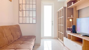 Una sala de estar con un sofá marrón, un televisor sobre un mueble de madera y una puerta que da a una habitación luminosa.